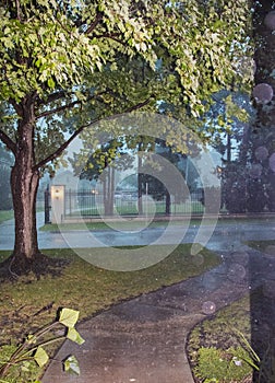 Looking out at street and fence and trees in urban neighborhood during a heavy rainstorm on stormy dark day with drops visible and