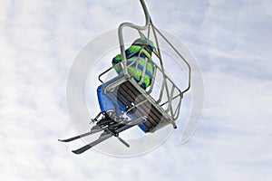 Looking up the skier in green and blue jacket, sitting on simple chairlift, sky with small clouds in background