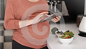 Looking up some recipes online. Cropped shot of a pregnant woman using her tablet while eating a salad in the kitchen.
