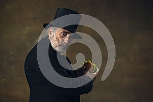 Handsome elderly gray-haired man, gentleman, aristocrat or actor eating fast food isolated on dark vintage background