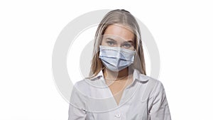 Looks sullenly young nurse in a medical mask and white uniform with blond straight hair looking at the camera. Isolated photo