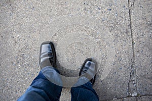 Loooking down at mans legs with blue jeans and black shoes on an urban city sidewalk walkway