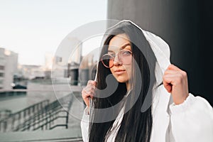 Ð¡lose-up portrait of attractive brunette in a white jacket and the sunglasses,standing against the backdrop of the city, looking