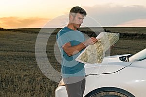 A lost man in the middle of the field with his car consults a map at sunset