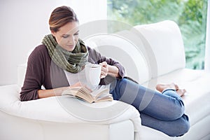 Lost in a page-turner...A young woman relaxing on her couch with a book and a cup of coffee.