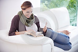 Lost in a page-turner...A young woman relaxing on her couch with a book and a cup of coffee.