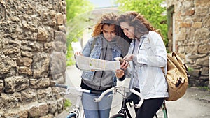 Lost tourists pretty girls are looking at paper map and smartphone screen standing in the street with bicycles and