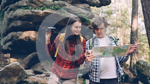 Lost tourists are standing in forest near huge rocks, looking at map then through binoculars and talking discussing way