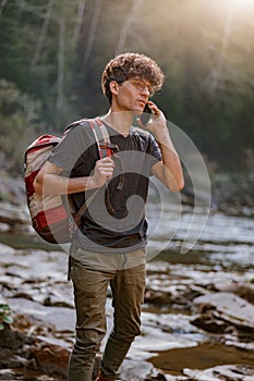 Lost young man tourist with backpack roaming at river in mountains and talking on cellphone.