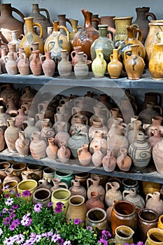 Lot of handicraft ceramic pots and vases on shelves and blooming plant flowers indoors. Crockery.