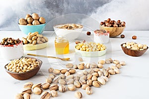 A lot of pistachio nuts are scattered on a white table. Wooden and ceramic bowls contain hazelnuts, almonds, walnuts, cashews.