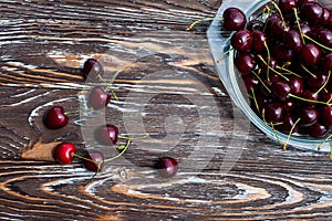 A lot of ripe red cherries on twigs lie in a glass plate on a dark blue wooden table with a striped texture.