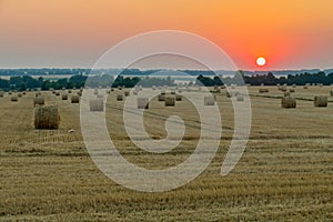 A lot of stacks of hay on the field under the sun setting in the horizon is red