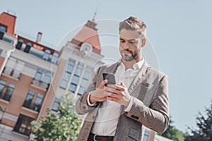 A lot to do. Serious brown-haired man is looking in his phone while standing in the park. He is going to work