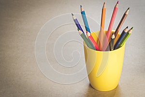 A lots of color pencils in yellow cup on wooden texture paper.