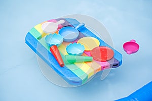 Lots of colorful pool toys laying on a multi colored swimming board floating on clear water in a small pool. Kids and playing in