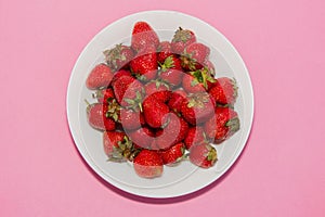 Lots of fresh red sweet strawberries on a white plate on a pink background.