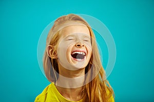 Loud and strong laughter of little girl with red hair, wearing yellow t-shirt, a shot of child on isolated blue.