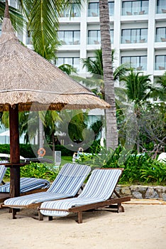 Lounge chair covered by beach umbrella thatched with palm leaves, multistory hotel background, coconut trees on white sandy shore