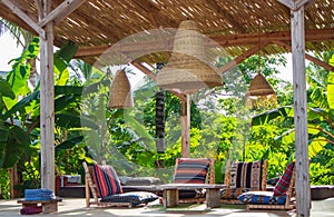 Lounge chairs with pillows and table under straw lamps in garden. Empty patio in tropical hotel resort.