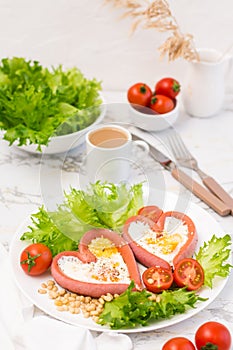 Love breakfast. Fried eggs in heart shaped sausages, lettuce and cherry tomatoes on a plate and a cup of coffee on the table.