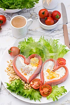 Love breakfast. Fried eggs in heart shaped sausages, lettuce and cherry tomatoes on a plate and a cup of coffee. Vertical view