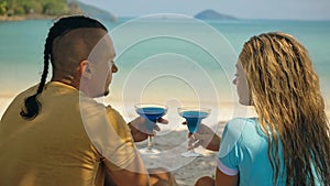 The love couple is holding a glass of blue curacao cocktail, on sea. Man with plait and blonde woman drink alcohol on