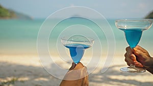 The love couple is holding a glass of blue curacao cocktail, on sea. Man and woman drink alcohol on sand beach. Summer