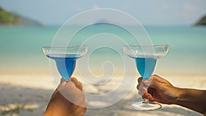 The love couple is holding a glass of blue curacao cocktail, on sea. Man and woman drink alcohol on sand beach. Summer