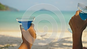 The love couple is holding a glass of blue curacao cocktail, on sea. Man and woman drink alcohol on sand beach. Summer