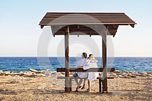 Love couple sitting on a bench by the sea and enjoying beautiful view at sunset