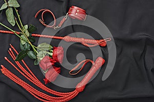 Love games. Top view of bdsm leather kit handcuffs, whip and beautiful red roses against of black silk fabric