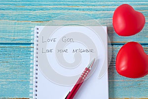 Love God and your neighbor handwritten quote in notebook with pen and red heart shapes on wooden background