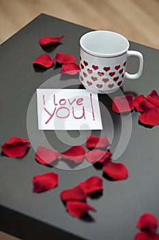 Love letter, rose petals and a cup on a dark coffee table.