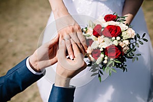 Love and marriage. Fine art rustic wedding ceremony outside. Groom putting golden ring on the bride`s finger. Bouquet of red and photo