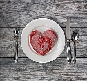 A love of meat, fresh ground beef in the shape of a heart on a white plate with cutlery and grey wood background