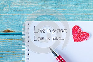 Love is patient and kind, handwritten verse in notebook with pen and red paper heart on wood