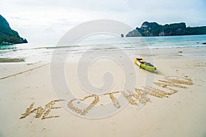 We love Thailand - text written by hand in sand on a sea beach with kayak over sky.