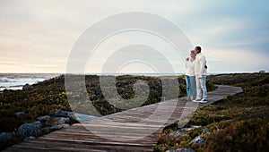 Love, travel and elderly couple hug on a boardwalk at a beach, calm and content against blue sky background. Senior, man