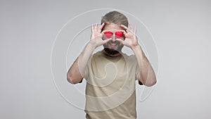 Love you! Romantic handsome bearded guy covering eyes with toy hearts, imitating heartbeat, playing having fun