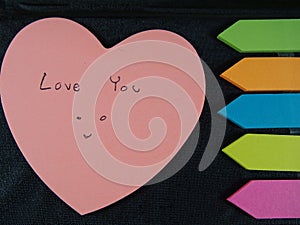 Love you with smiling face, drawing and writing on pose its paper with colorful heart and arrow on black leatherette background