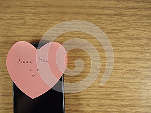 Love you with smiling face writing on sticker note and black mobile phone on wood background