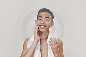 Love your skin. Portrait of shirtless young asian man with towel around his neck looking at camera, applying lotion