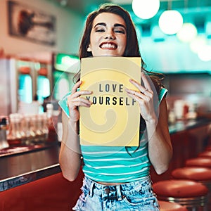 Love yourself. Cropped portrait of an attractive young woman holding up a sign in a retro diner.