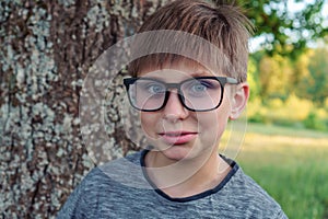 Loveable boy with blue eyes, in glasses in gray longsleeve standing in front of tree in park and enjoying fresh air.