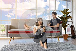 Lovely Asian couple in living room at home. young woman with headphone sit on floor playing video game, man lying on couch working