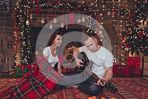 Lovely beautiful couple woman and man are sitting at home near fireplace and playing guitar. Christmas time.