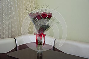 Lovely big bouquet with flowers of roses of vinous red color are standing in the glass vase on the table. Green leaves and thorns.