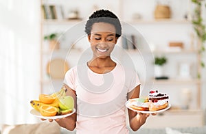 Lovely black lady holding plates with fruits and sweets, looking at unhealthy pastries, tempted to eat them at home