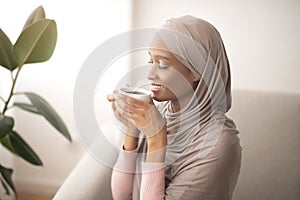 Lovely black lady in traditional hijab smelling coffee, enjoying magical aroma with closed eyes, indoors